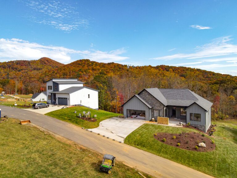 A scenic aerial view of a modern, two-story house nestled in a lush, autumnal landscape. The house features a white exterior, a garage, and a covered patio area. The surrounding hills are blanketed in vibrant fall foliage, creating a picturesque rural setting. A small utility vehicle is visible on the driveway, adding to the tranquil, countryside atmosphere.