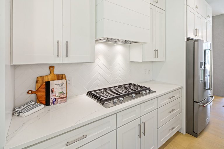 A modern, well-equipped kitchen with sleek white cabinetry, a gas stovetop, and a marble-like countertop. A wooden cutting board and a cookbook add a touch of warmth and functionality to the space, while the herringbone-patterned backsplash adds visual interest and texture.