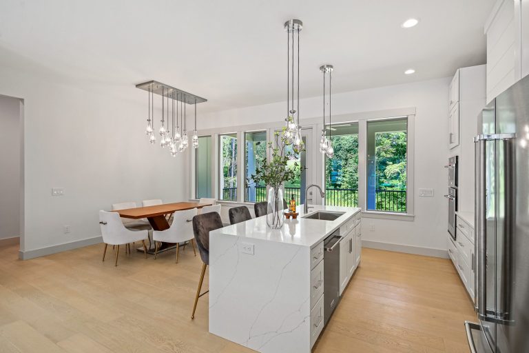 A modern, open-concept kitchen and dining area with a large marble island, sleek pendant lighting, and floor-to-ceiling windows overlooking a lush, natural landscape outside. The space features a mix of contemporary and natural elements, creating a bright and inviting atmosphere.