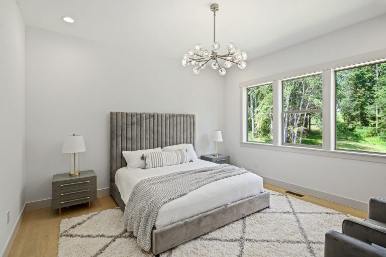 A modern, well-appointed bedroom with a plush, gray upholstered headboard, a large crystal chandelier, and floor-to-ceiling windows overlooking a lush, green landscape. The room features a cozy, neutral color palette and comfortable bedding, creating a serene and inviting atmosphere.