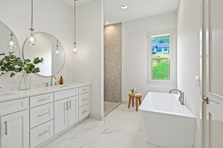 A modern and spacious bathroom with a large vanity, circular mirrors, and pendant lighting. The room features a freestanding bathtub, a tiled shower, and a window overlooking a lush, green landscape outside. The overall design creates a serene and spa-like atmosphere.