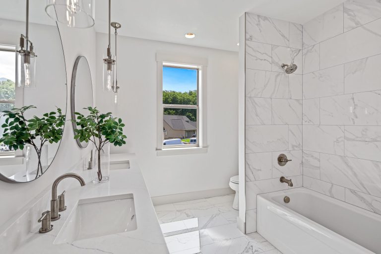 A modern, bright bathroom with sleek marble tile walls, a large vanity mirror, and a view of the lush greenery outside through a window. The space features contemporary fixtures, including a glass-enclosed shower, a freestanding bathtub, and a stylish sink with a chrome faucet.