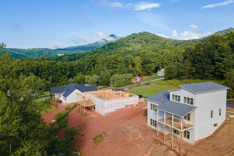 Aerial view of a residential construction site nestled in a lush, verdant landscape. The image shows multiple buildings in various stages of construction, surrounded by rolling hills, dense forests, and a clear blue sky with wispy clouds. The scene conveys a tranquil, natural setting amidst the ongoing development.