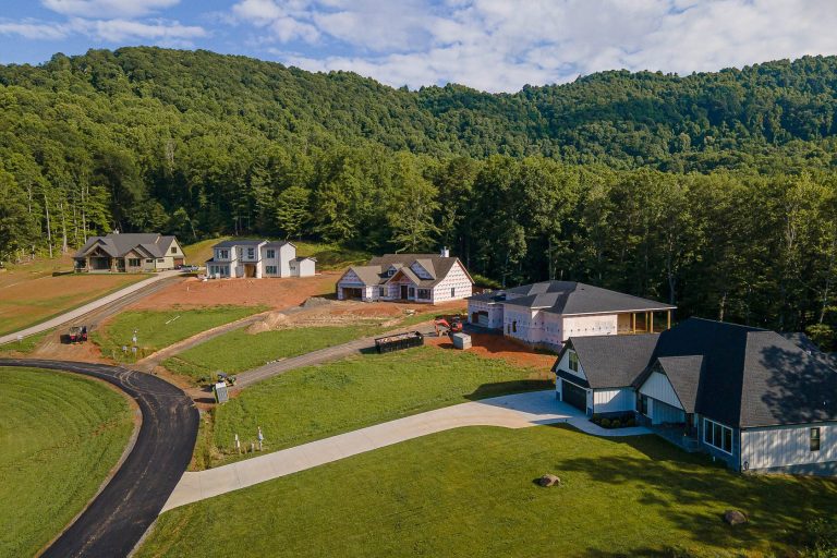 Aerial view of a residential neighborhood nestled in a lush, forested landscape. Several newly constructed homes with varying architectural styles are situated on gently sloping terrain, surrounded by a network of paved roads and green lawns. The dense, verdant trees and hills in the background provide a picturesque natural backdrop, creating a serene and picturesque suburban setting.