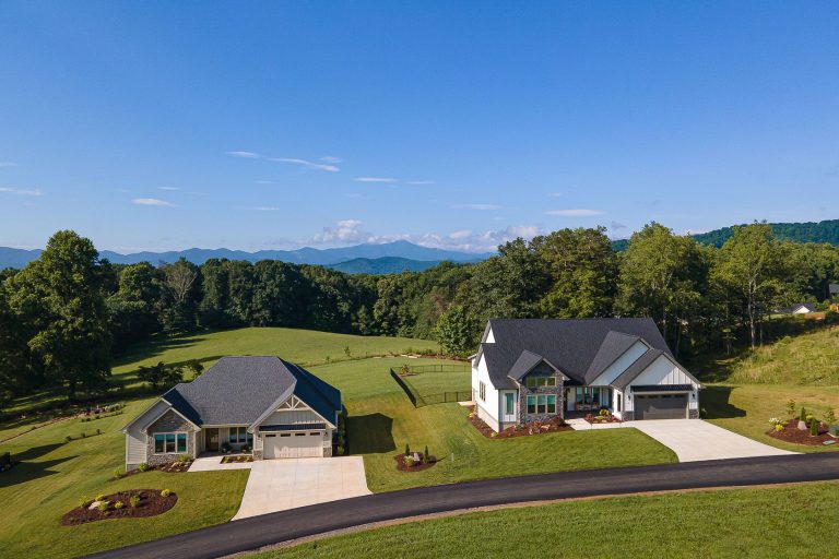 A large, modern two-story house with a garage nestled in a lush, green landscape. The house is surrounded by a well-manicured lawn, mature trees, and distant mountains can be seen in the background, creating a picturesque, serene setting.
