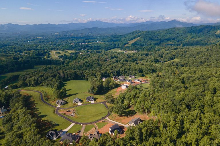 Aerial view of a lush, forested mountain landscape with a residential development nestled among the trees. The image showcases rolling hills, winding roads, and a mix of homes and natural surroundings, creating a picturesque rural setting.