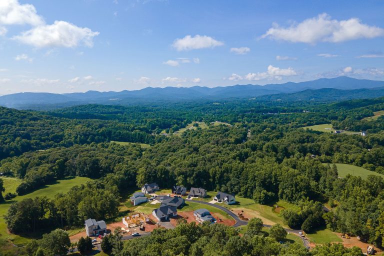 Aerial view of a lush, green mountain landscape with a residential development nestled among the trees. The image showcases rolling hills, dense forests, and distant blue mountains, creating a picturesque and serene natural setting.