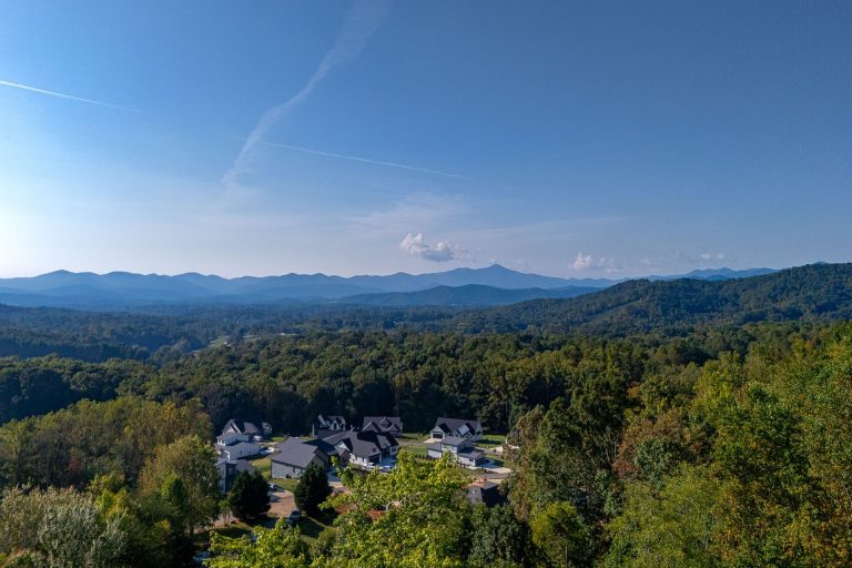 A scenic aerial view of a residential community nestled among lush, rolling hills and mountains. The landscape is dotted with densely forested areas, interspersed with clusters of homes with dark roofs. In the distance, the silhouettes of distant mountain peaks can be seen against a clear blue sky with wispy clouds.