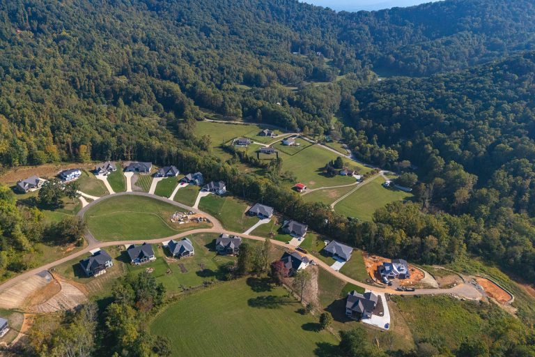 Aerial view of a residential development nestled in a lush, forested landscape. The image shows a series of houses arranged in a curved, circular pattern, surrounded by expansive green lawns and trees. In the background, rolling hills and mountains create a picturesque natural setting, adding to the serene and tranquil atmosphere of the scene.
