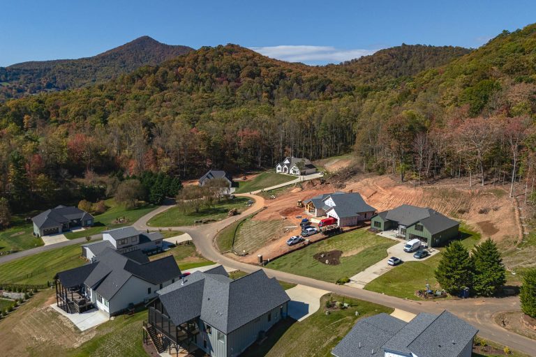 Aerial view of a residential community nestled in a picturesque mountain landscape. The image showcases a collection of modern homes with slate roofs, surrounded by lush, colorful foliage and rolling hills. A winding road leads through the community, with construction vehicles visible, indicating ongoing development. The scenic backdrop of the mountains and forests creates a serene and peaceful setting, highlighting the natural beauty of the area.