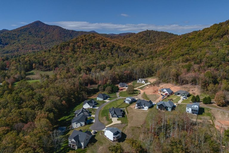 Aerial view of a residential neighborhood nestled in a lush, mountainous landscape. The homes are surrounded by vibrant autumn foliage, with a winding road leading through the community. The rolling hills and dense forest create a picturesque, serene setting, showcasing the natural beauty of the region.