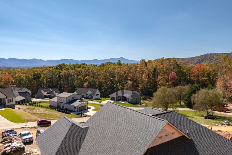 Aerial view of a residential neighborhood nestled in a picturesque autumn landscape. The image showcases a community of homes surrounded by a lush, colorful forest with vibrant fall foliage. The homes have dark-colored roofs and are situated on well-manicured lawns, creating a serene and picturesque setting against the backdrop of the rolling mountains in the distance.